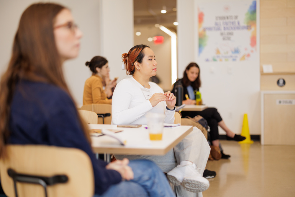 People sitting at tables in a classroom, attentively listening. A sign with text is visible on the wall in the background.