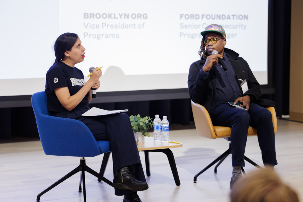 Two people sit on stage holding microphones in front of a screen displaying text about Brooklyn.org and Ford Foundation roles.