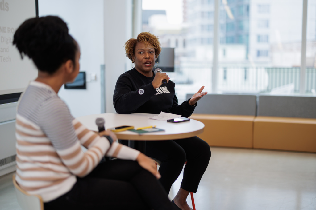 Two people are sitting at a table, engaging in a discussion. One is speaking into a microphone, while the other listens attentively. They are indoors with a window in the background.