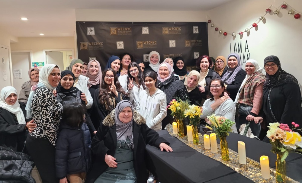 A group of women and girls, some wearing headscarves, gather in a room. A table with flowers and candles is in front of them. A banner and wall decor are in the background.