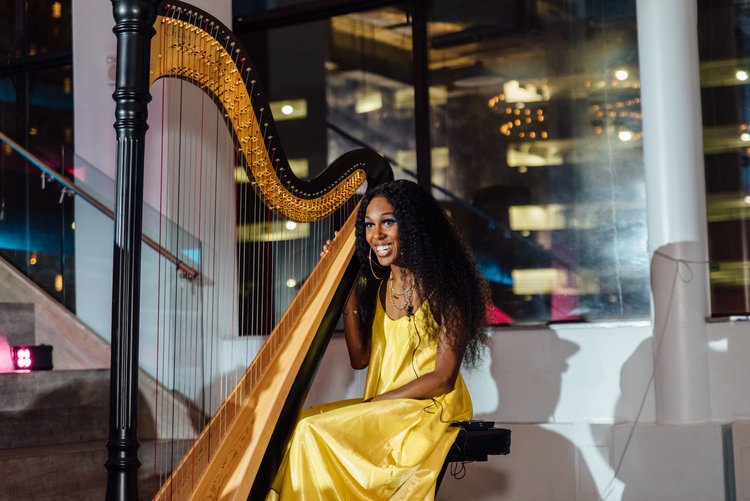 Person in a yellow dress playing a harp indoors near stairs and a window.