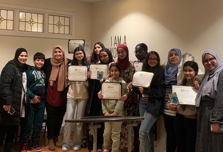 A group of people, including children and adults, standing indoors. Several individuals are holding certificates.