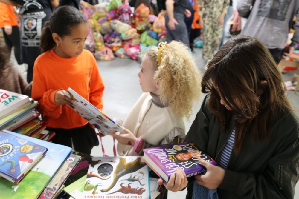 Three children browse and discuss books at a book fair, surrounded by stacks of colorful children's books.