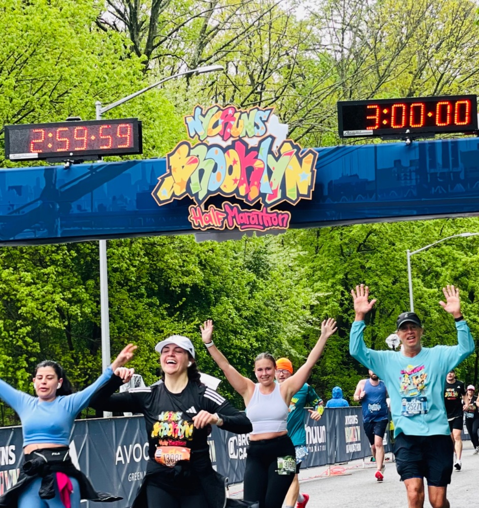 Runners celebrate as they cross a marathon finish line, surrounded by trees. A clock displays a time of 3:00:00.