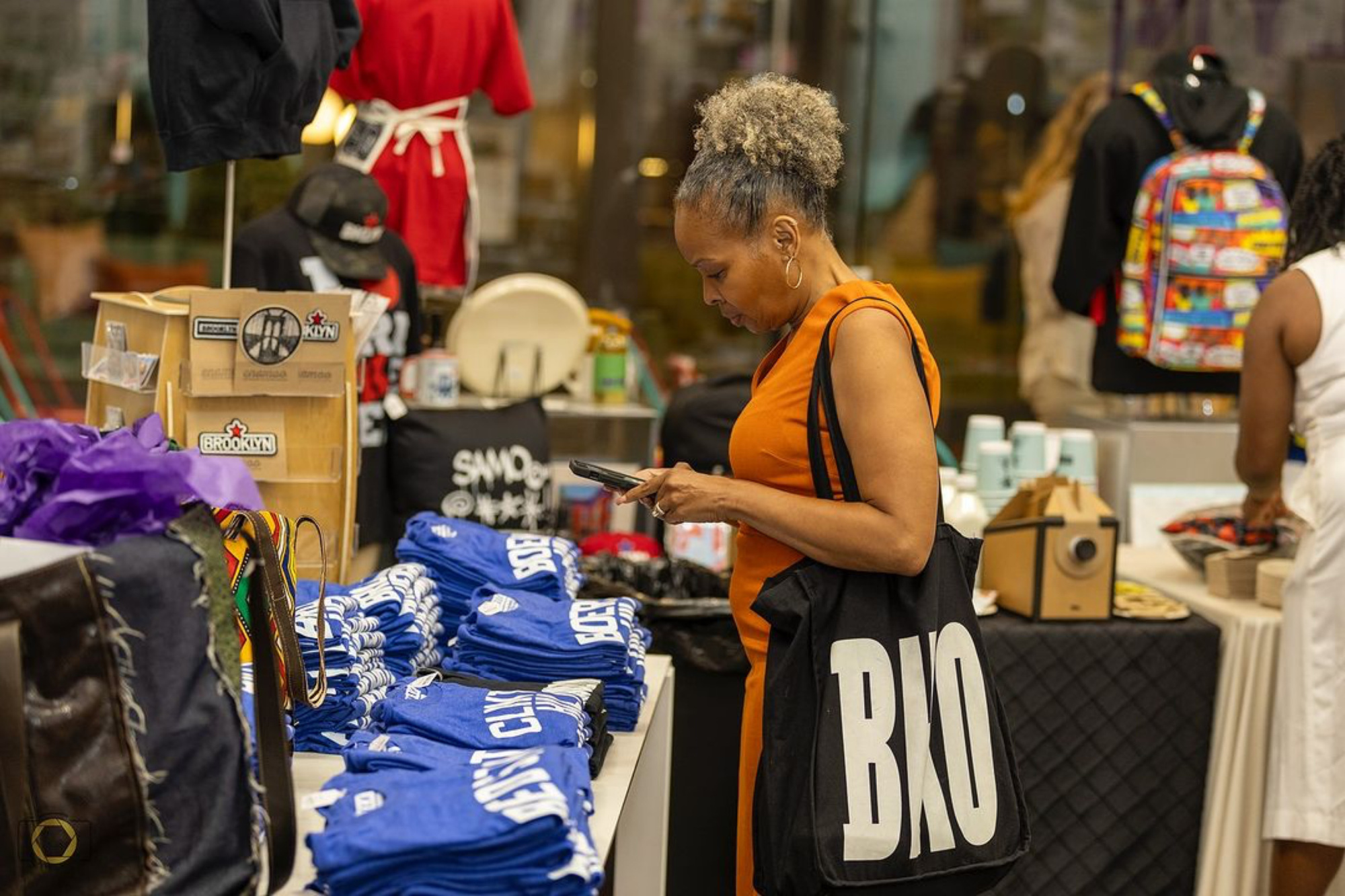 A woman in an orange dress looks at her phone while shopping at a vendor stall featuring blue shirts and various merchandise. She carries a black tote bag with "BRO" printed on it.