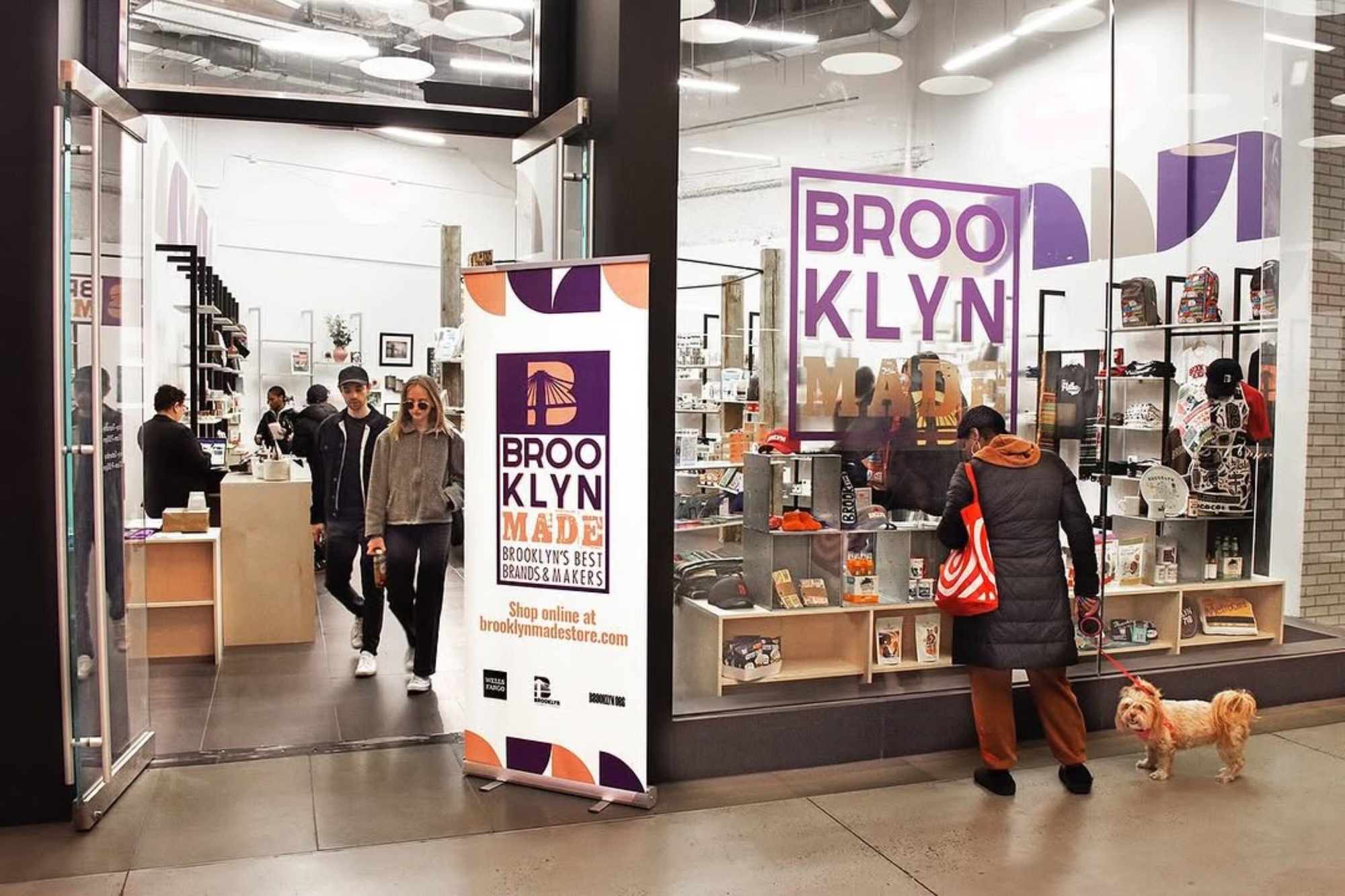 People walking by and entering a store named "Brooklyn Made," displaying products in the window. A woman with a small dog is looking at the display.