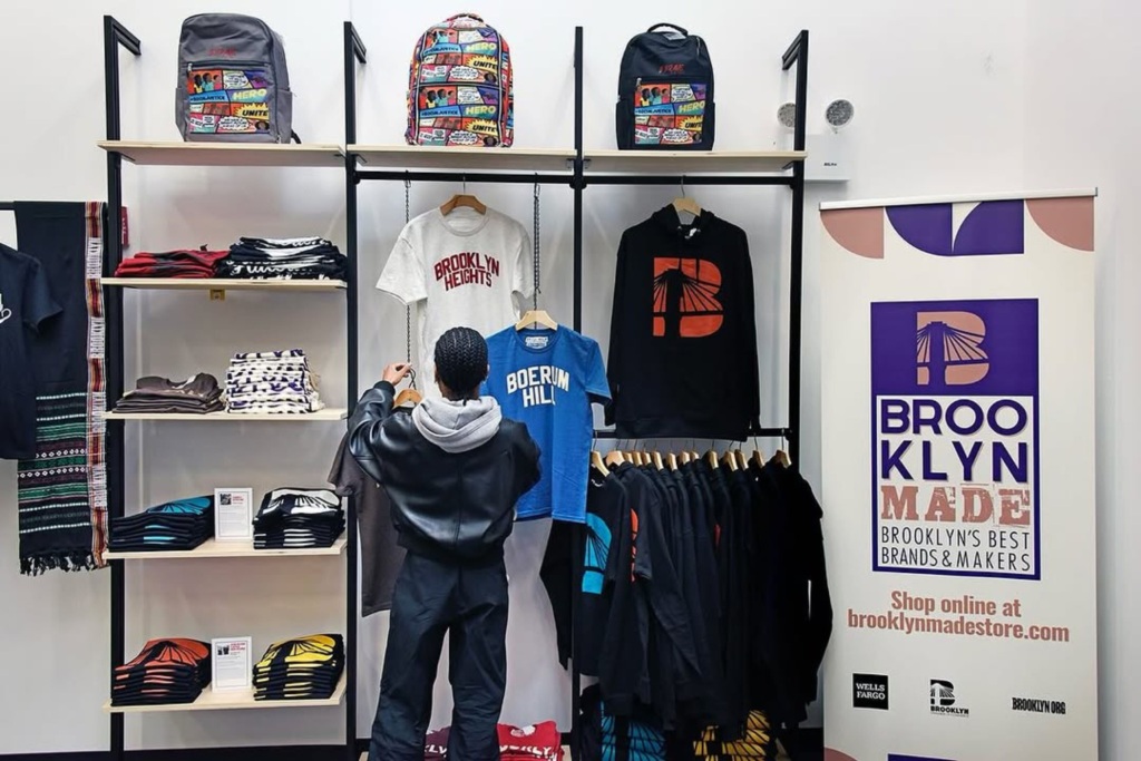 Person browsing clothing and accessories in a store. Shelves display shirts, hoodies, and backpacks. A sign reads "Brooklyn Made: Brooklyn's Best Brands & Makers.