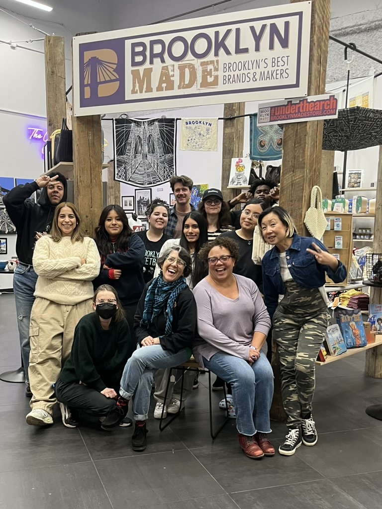 A group of people pose together under a "Brooklyn Made" sign in a store. Some are sitting, while others stand behind them. Shelves with various items are visible in the background.