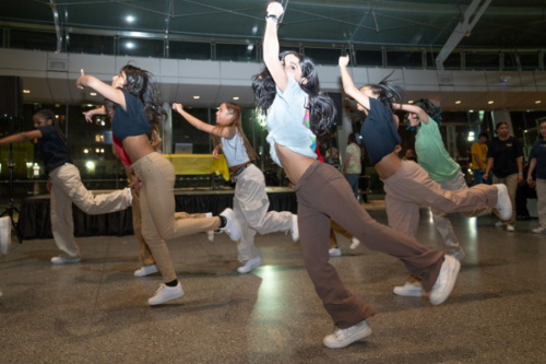 A group of dancers energetically performing in a well-lit indoor space, all in coordinated casual attire and sneakers.