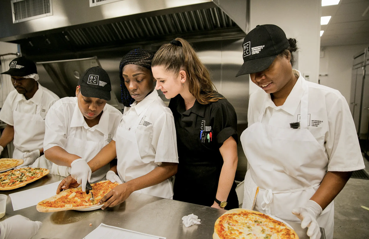 People in a kitchen prepare pizzas, wearing uniforms and hats. One person is instructing others while they work.