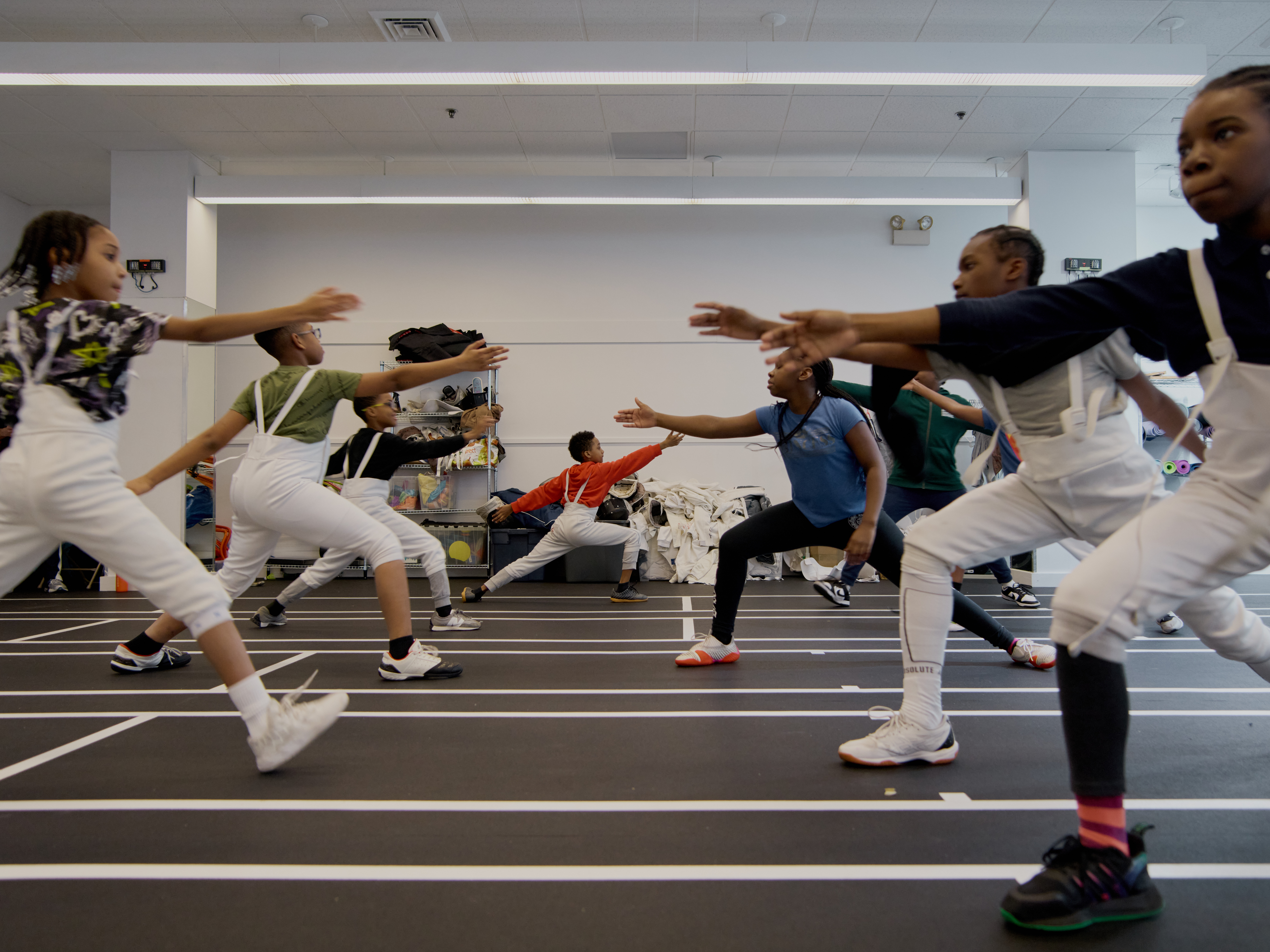 People practicing fencing footwork in a room, wearing protective gear and holding fencing weapons, performing lunges in a group setting.