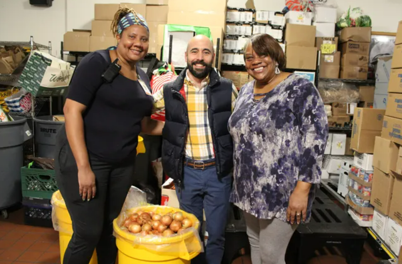 Three people stand in a storeroom filled with boxes and bags. They are smiling and posing beside a yellow bin filled with onions.