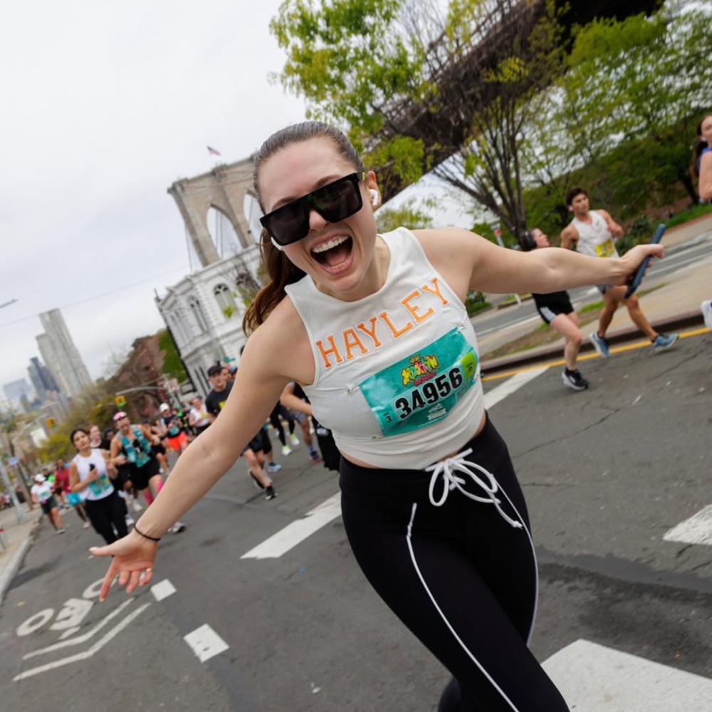 A woman in athletic clothing runs with outstretched arms on a city street, wearing a race bib numbered 34956. Buildings and a bridge are visible in the background.