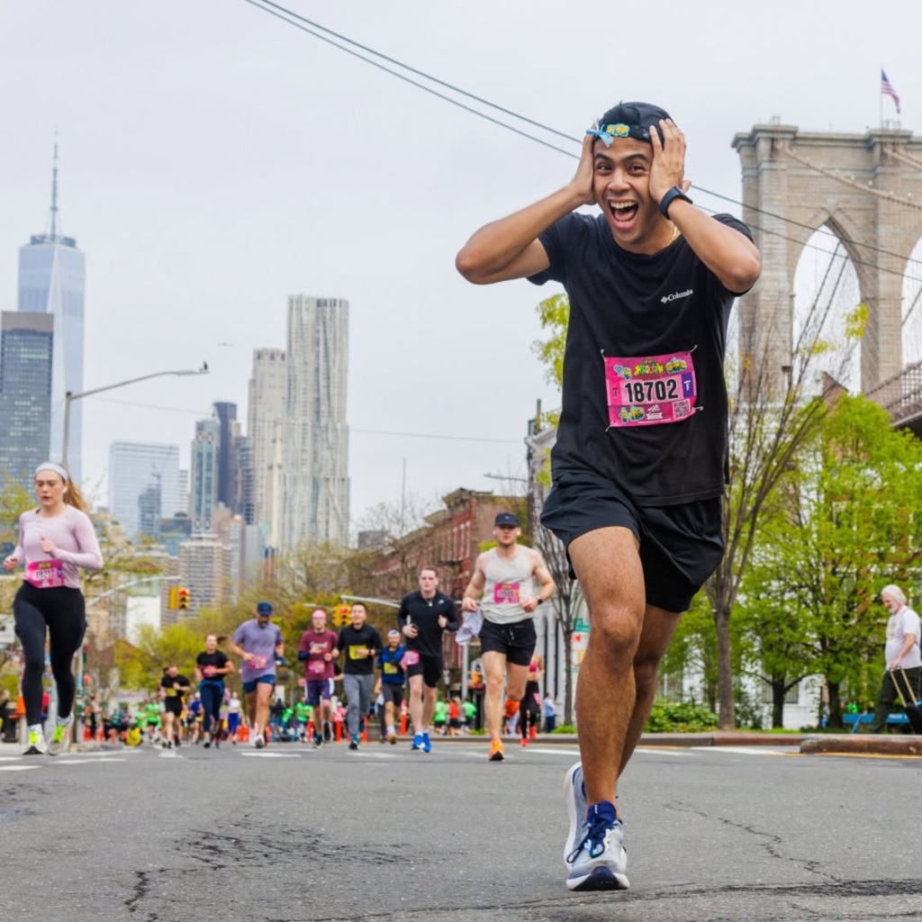 A runner in black sportswear expresses excitement during a race with the city skyline and a bridge in the background.