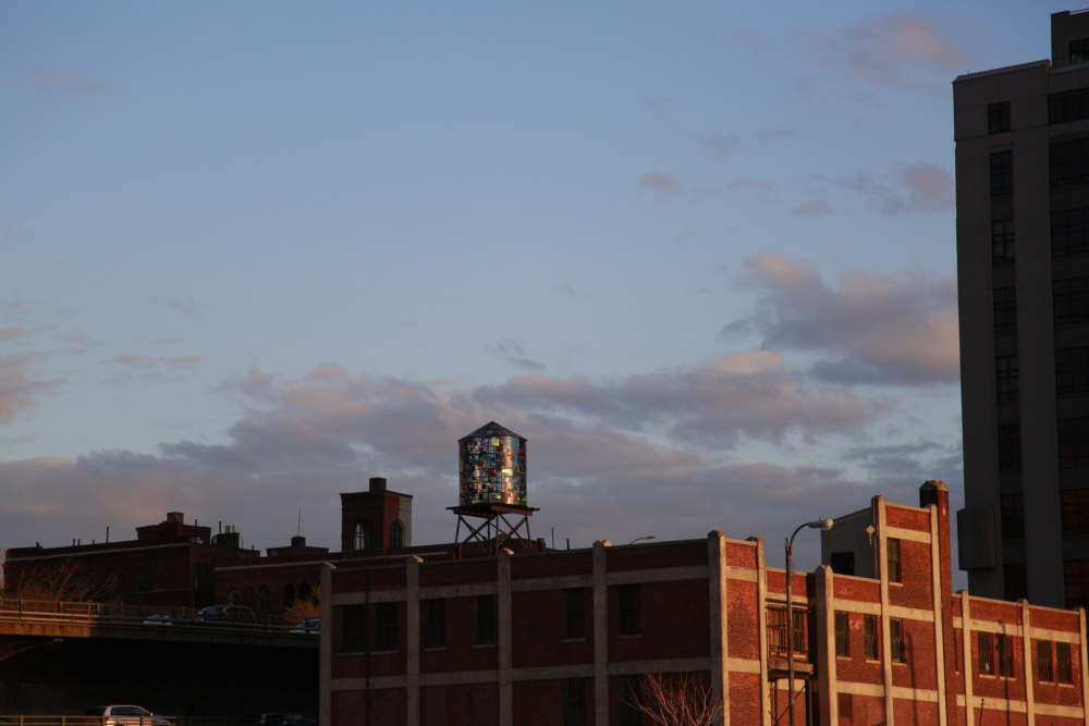 A cityscape at dusk with a brick building in the foreground and a colorful water tower on the roof. The sky is partly cloudy.