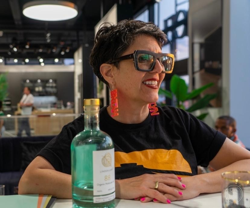 Person wearing large sunglasses and red earrings, smiling at a table with a bottle in front, inside a modern, well-lit space.