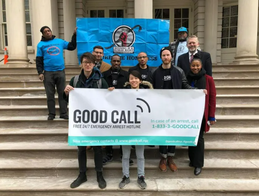 A group of people stands on steps holding a banner that says "GOOD CALL: Free 24/7 emergency arrest hotline" with a phone number. A sign about homelessness is on display behind them.