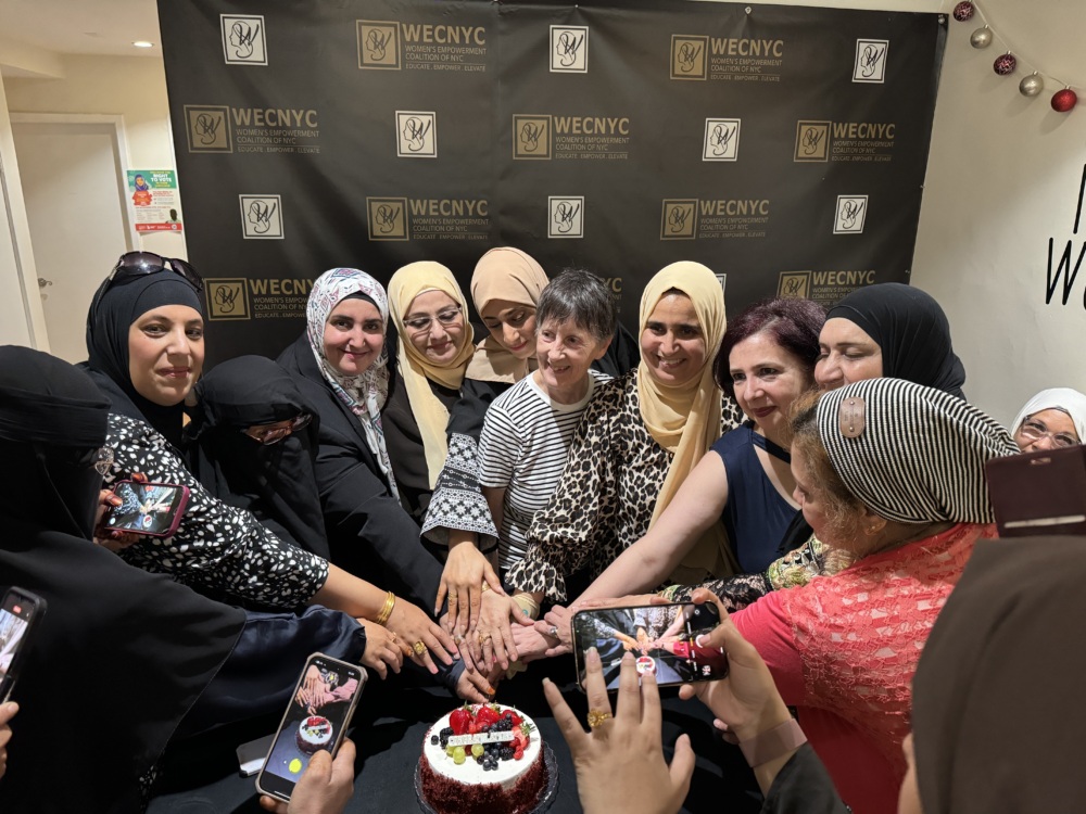 A group of women gathered around a small cake, touching hands, in front of a WECNYC backdrop; some attendees photograph the scene.