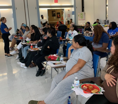 People seated in a bright, spacious room, attending an educational event. They are listening attentively, with notebooks and snacks on tables.