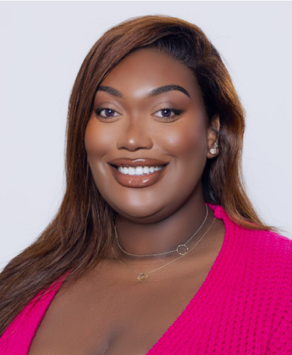 Woman with long brown hair smiles, wearing a bright pink top and layered necklaces against a plain background.