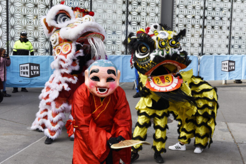 Performers in colorful dragon costumes dance at an outdoor event, with a crowd and a security officer in the background.