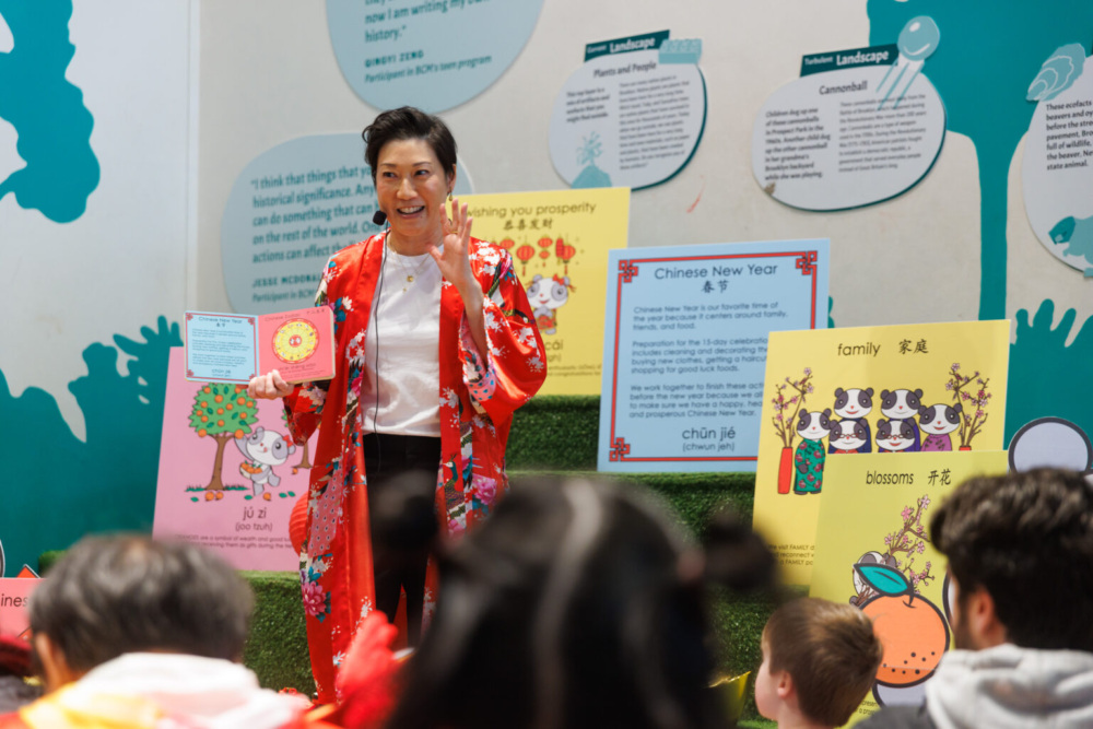 A woman in a red outfit talks to children at a Chinese New Year event, with colorful posters and decorations in the background.