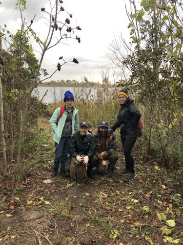 Four people in outdoor gear pose in a wooded area near a body of water. Two are standing, and two are crouching, surrounded by trees and foliage under an overcast sky.