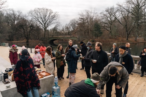 A group of people gathered outdoors in a park, wearing winter clothing. Some are interacting and holding cups near a stone barrier and a body of water. Trees and a bridge are visible in the background.