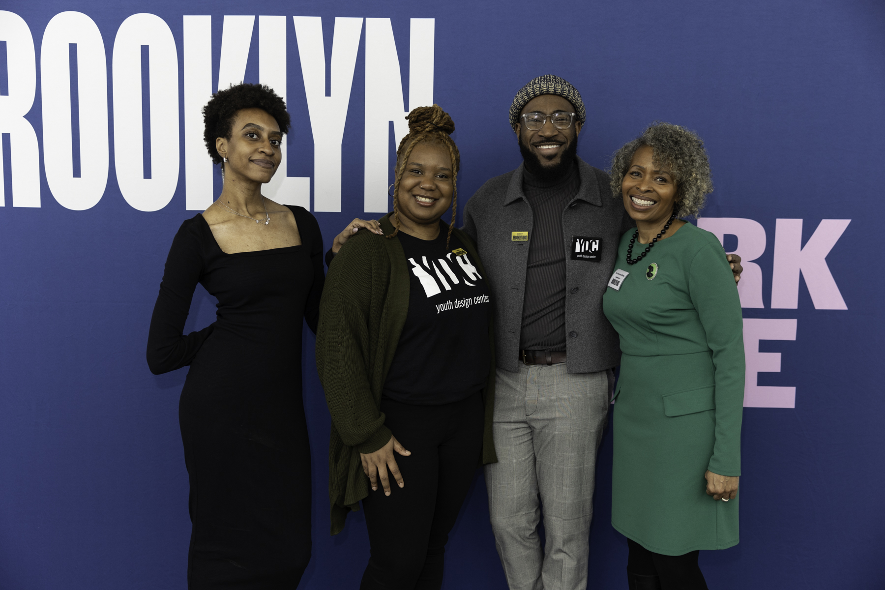 Four people standing in front of a blue background with "Brooklyn" written on it, smiling and posing for the photo.