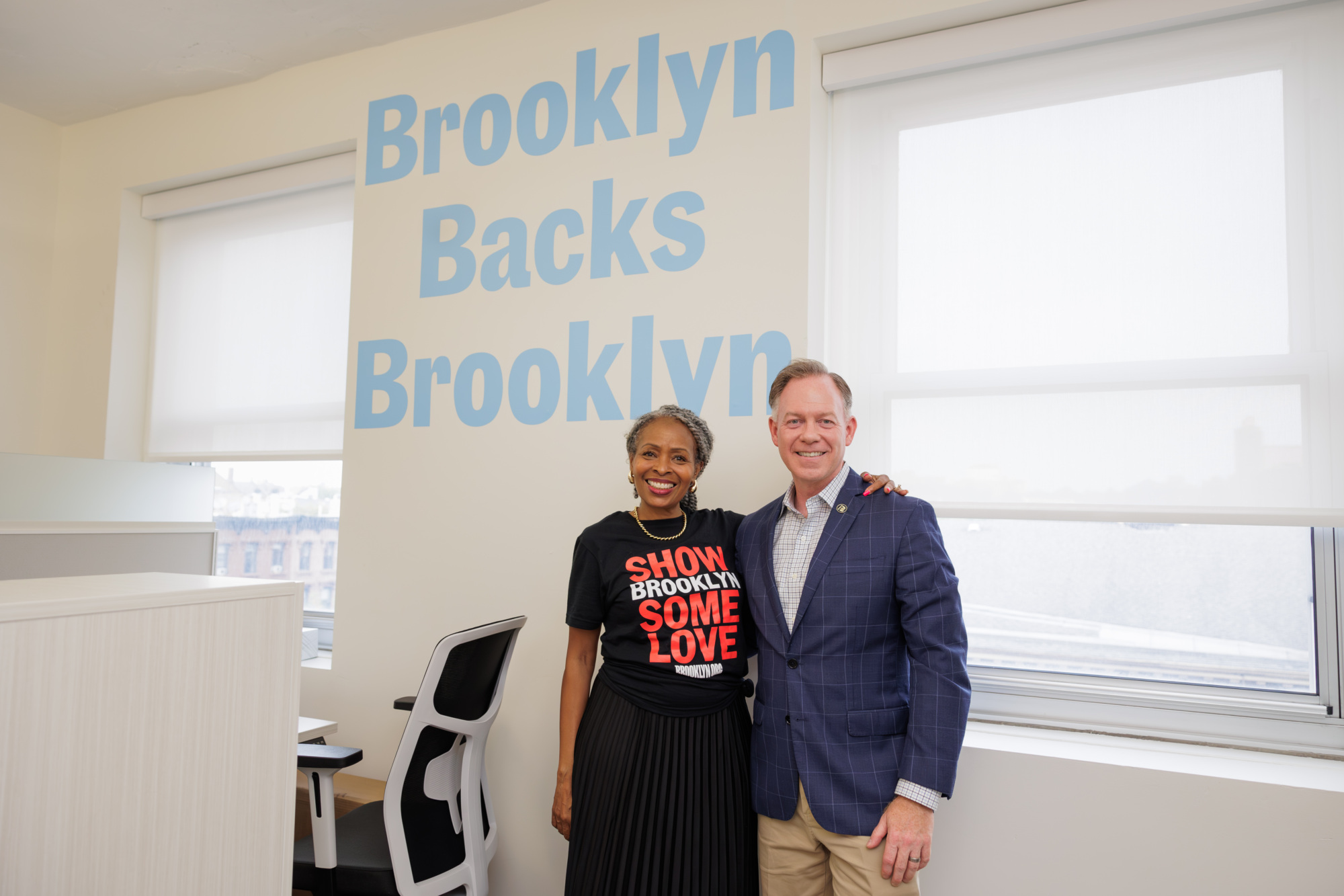 A woman and a man stand smiling in an office with "Brooklyn Backs Brooklyn" written on the wall behind them.