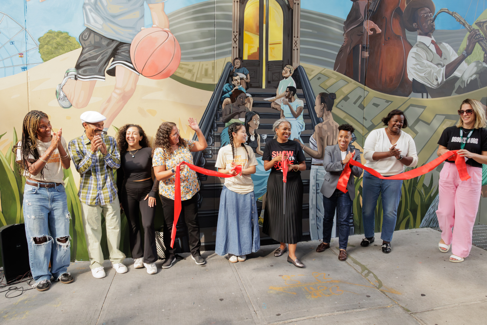 A group of people stand in front of a mural, holding and cutting a red ribbon in a ribbon-cutting ceremony.