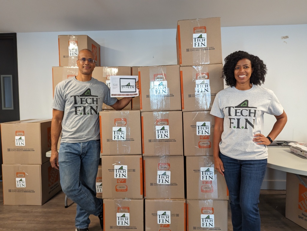 Two people wearing "Tech F.I.N." shirts stand next to stacked boxes with the same logo in an office setting.