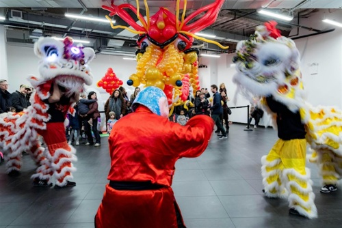 Lion dancers in colorful costumes perform in a room with an audience watching. A large decorative balloon dragon is in the background.