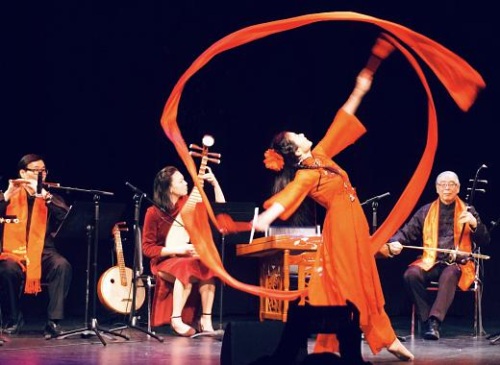 A dancer in flowing red performs with a red ribbon on a stage, accompanied by musicians playing traditional instruments.