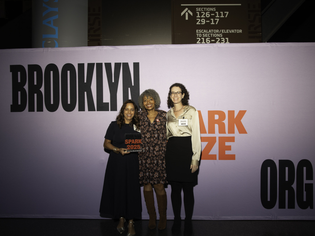 Three women stand in front of a backdrop with the words "Brooklyn" and "Spark Prize." One woman holds a small award.