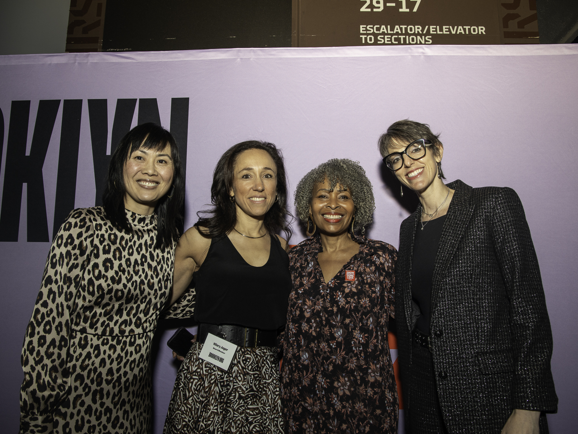 Four people stand smiling in front of a purple background with partial text, under a sign indicating escalators or elevators.