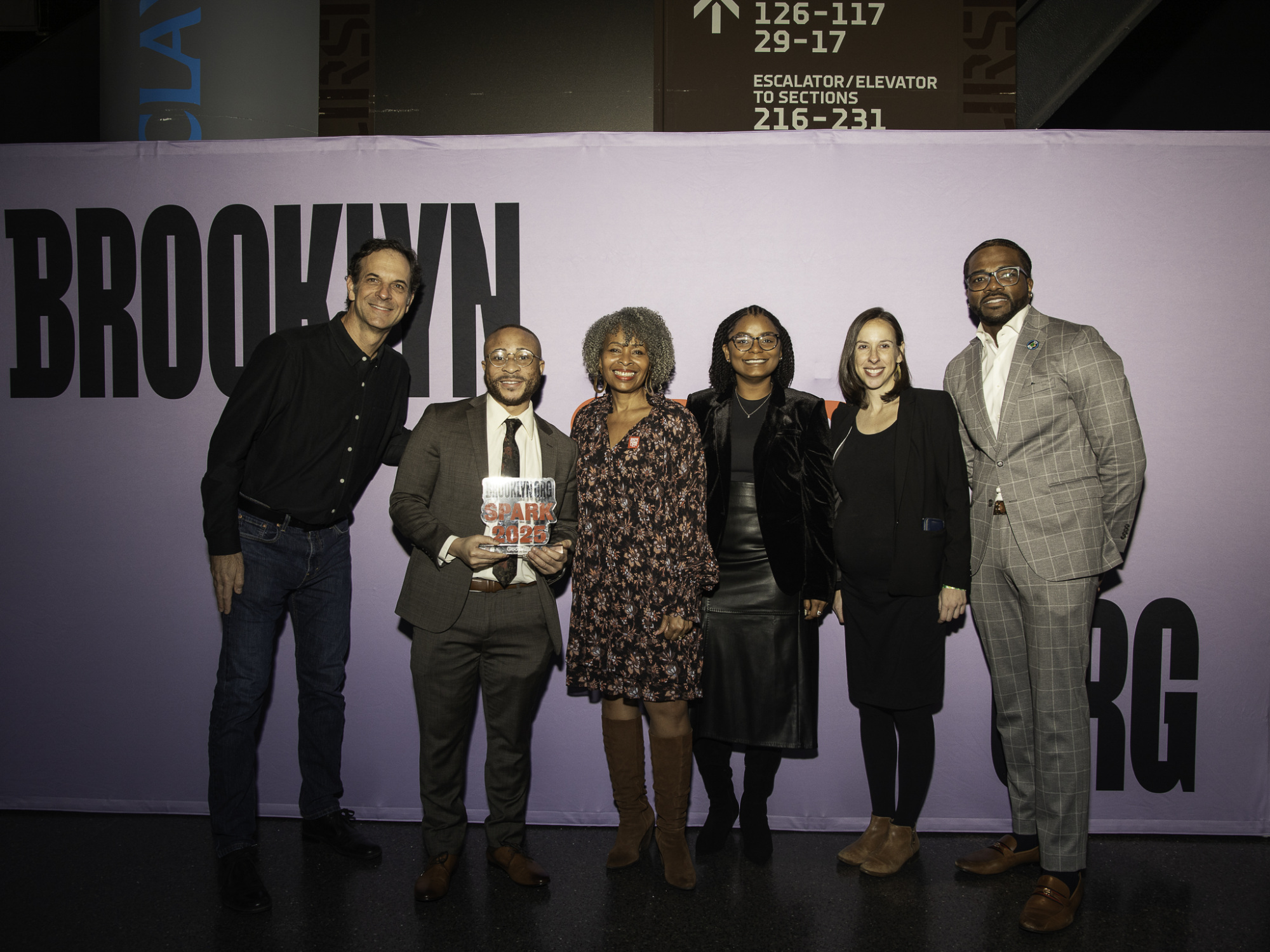 Six people standing in front of a purple Brooklyn sign, with one person holding a book. They are dressed in formal attire.