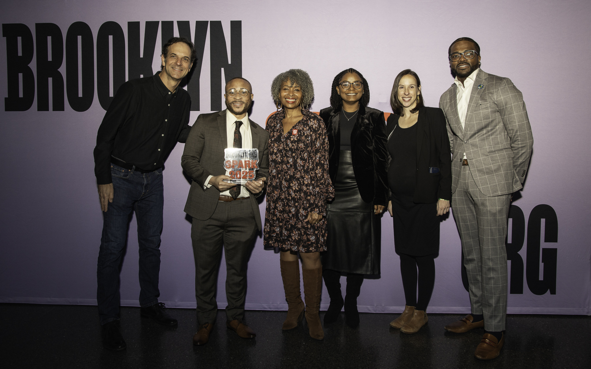 Six people standing in front of a purple Brooklyn sign, with one person holding a book. They are dressed in formal attire.
