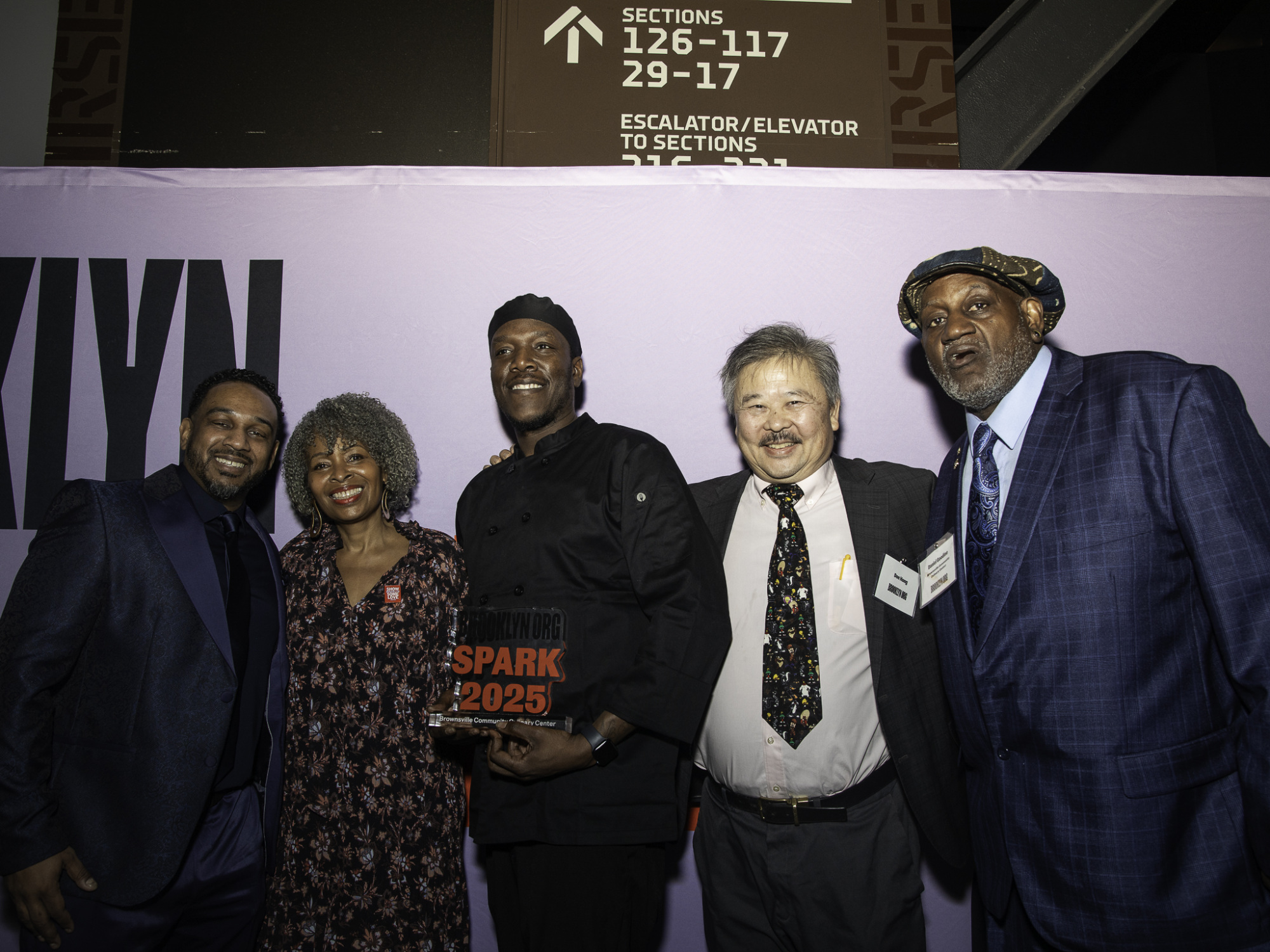 Five people standing together, one holding a "Spark 2025" sign, in front of a backdrop with directional signage.