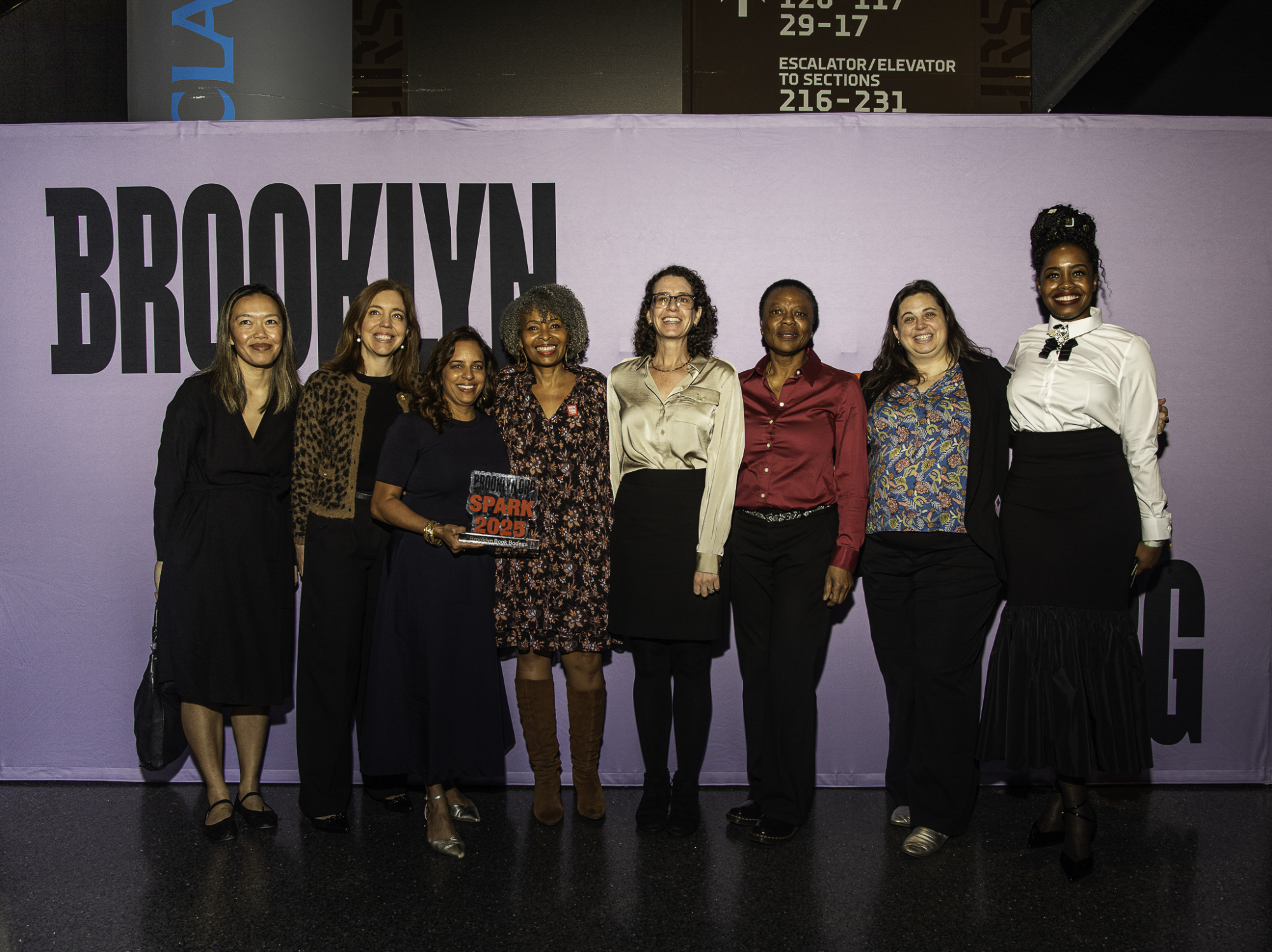 Eight people standing in front of a backdrop with "BROOKLYN" visible, one is holding a "SPARK 2023" award.