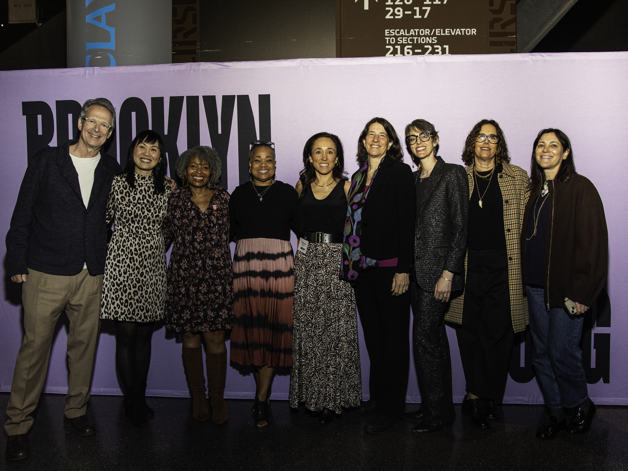 A group of eight people standing in front of a purple backdrop with partial text "PROXIMITY" visible. They are smiling and wearing a variety of outfits.