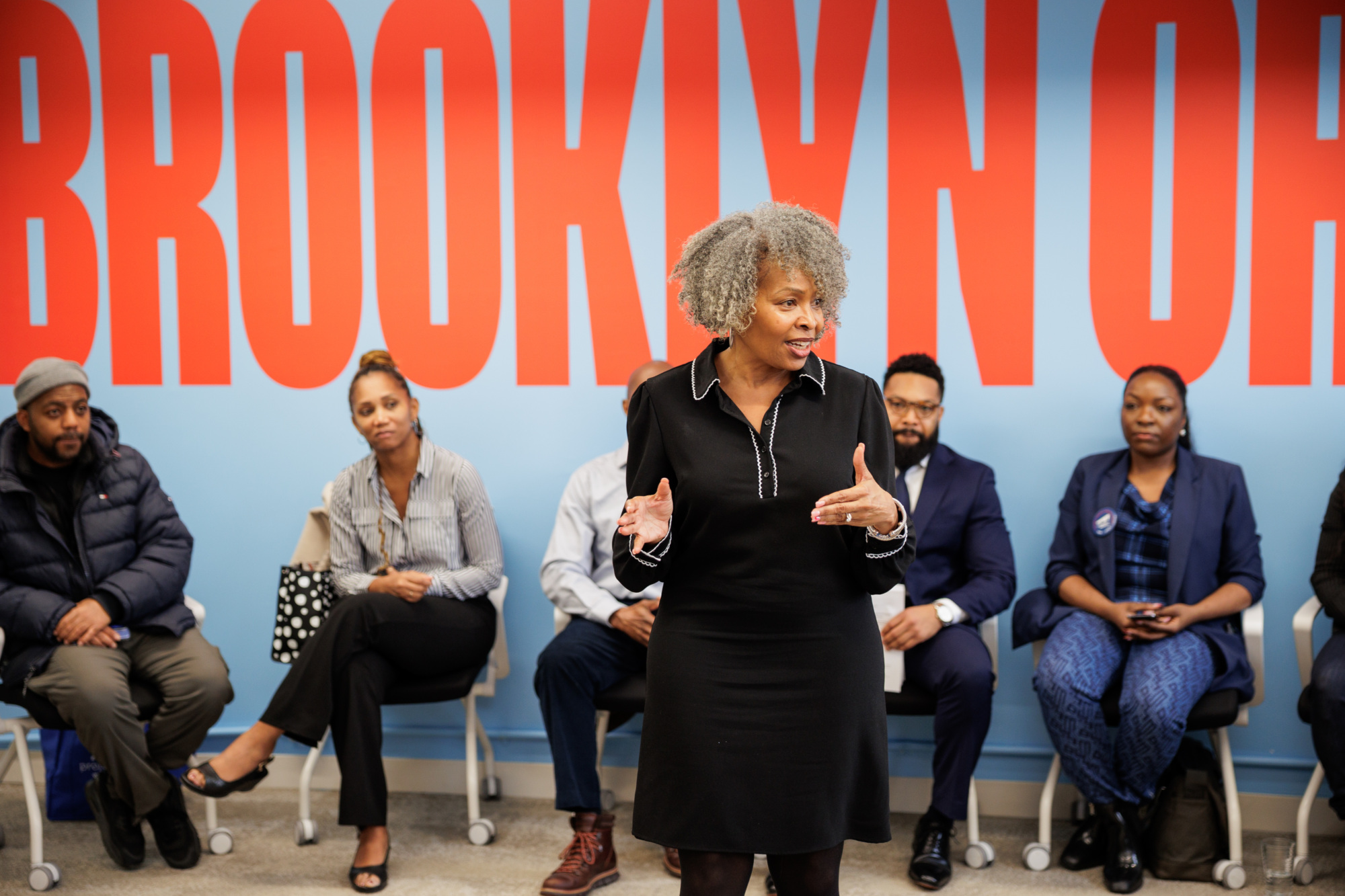 A woman speaks to an audience seated in chairs, with large red letters spelling "BROOKLYN OH" on the blue wall behind them.