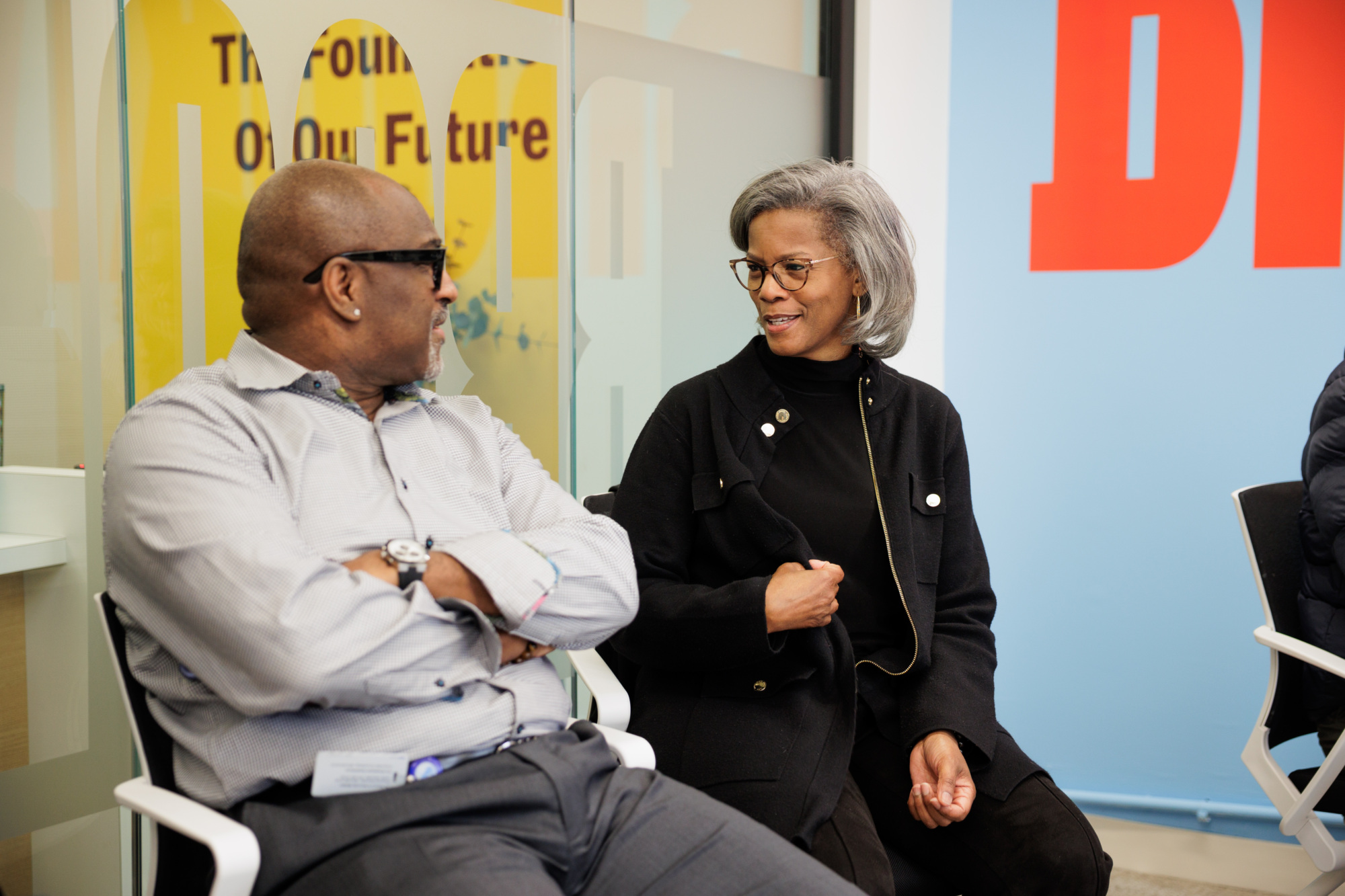 A man and a woman are seated in a room, engaged in conversation. They are wearing glasses and business attire. Behind them, a wall displays partial text and colorful graphics.