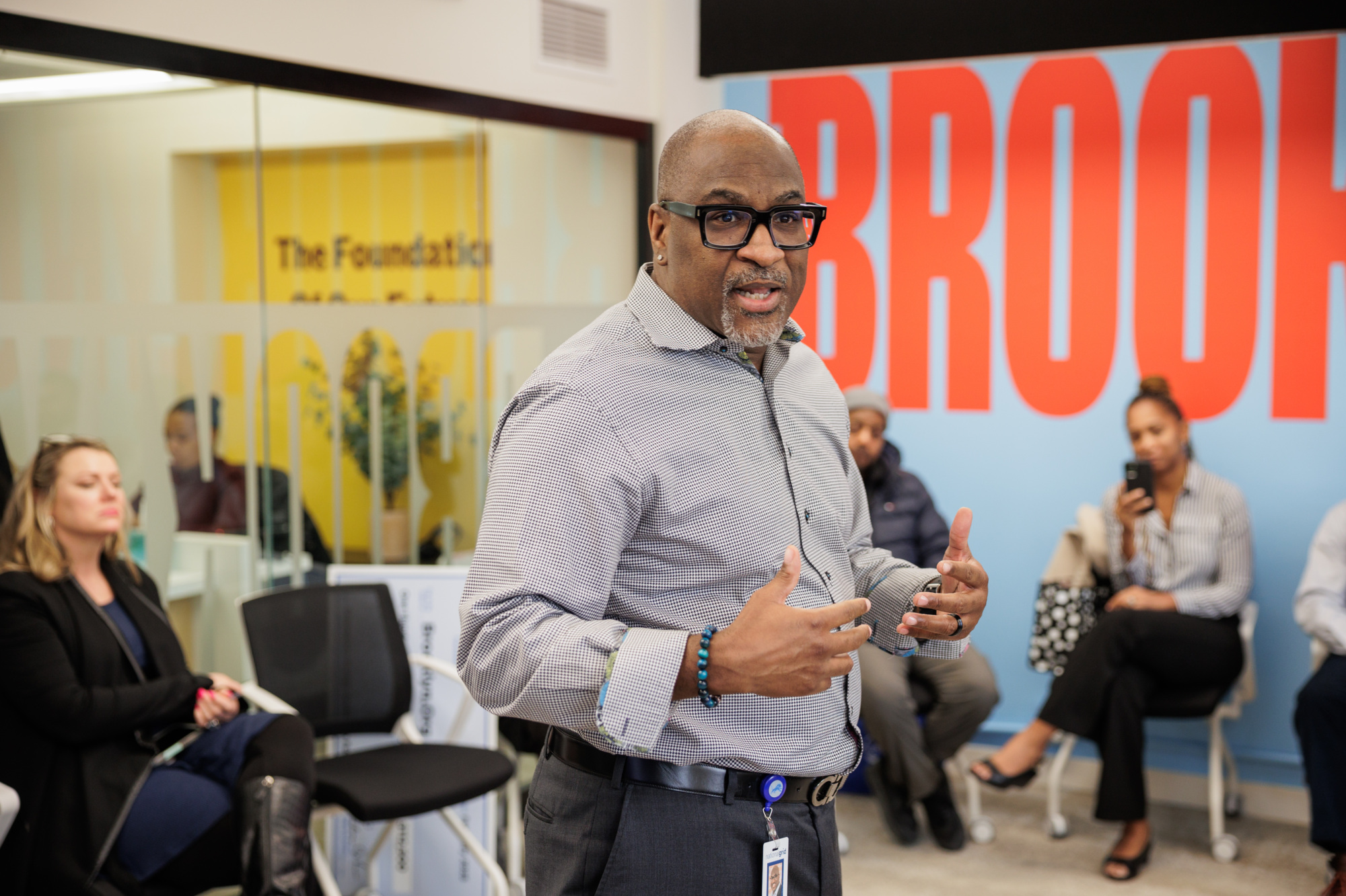 A person speaking in a room with seated individuals in a discussion setting. A large red word "BROOK" is partially visible on a wall behind.