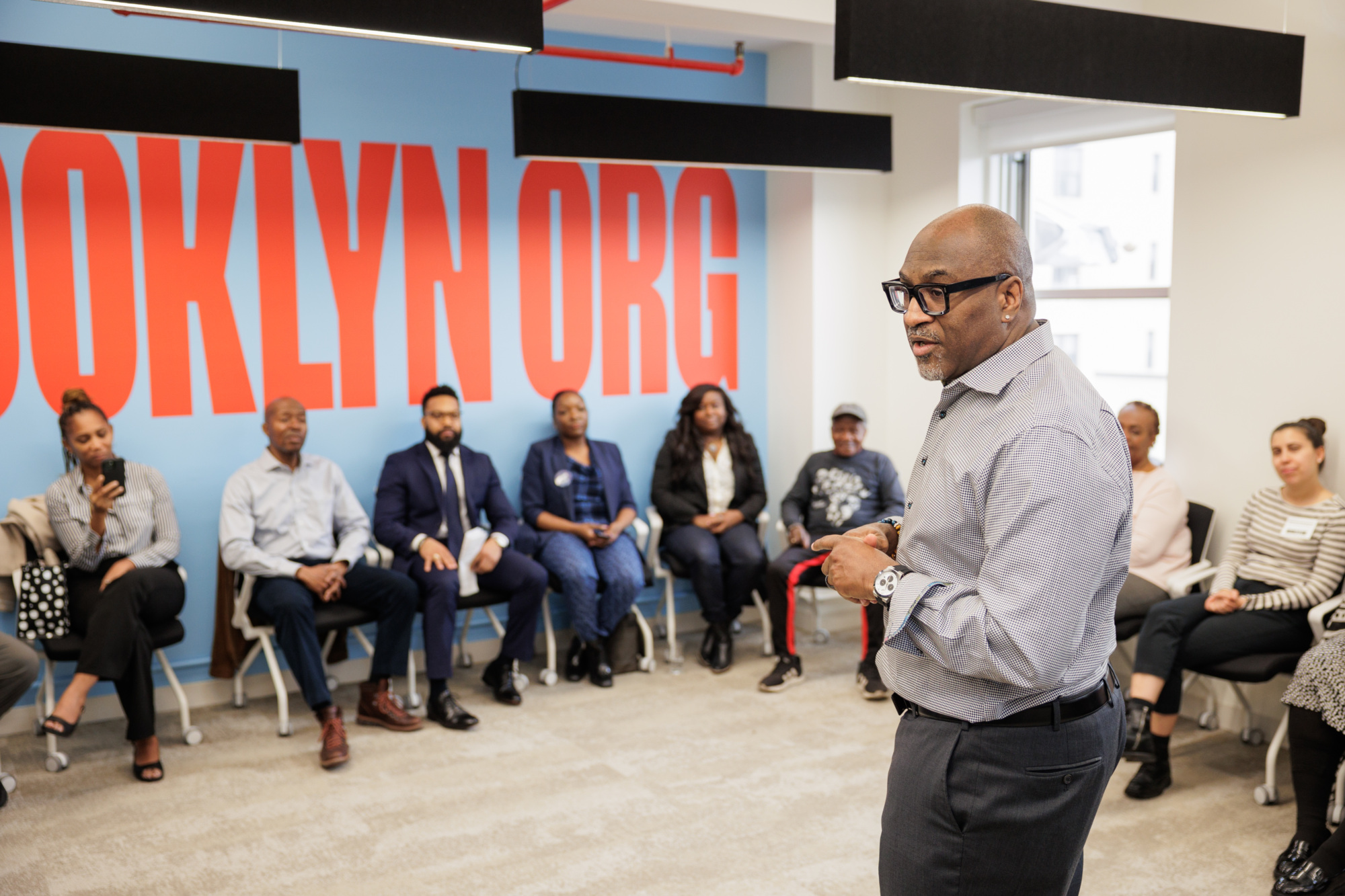 A man speaks to a group of seated people in a room with "BROOKLYN ORG" on the wall.