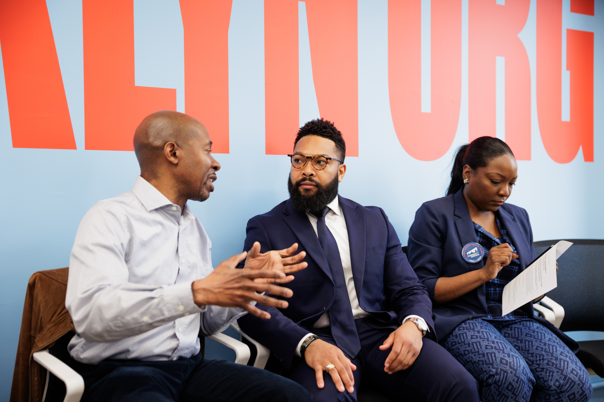 Three people seated and talking; two men in conversation, one wearing a suit, beard, glasses. A woman in a blazer looks at a phone. Background has large red text on a blue wall.