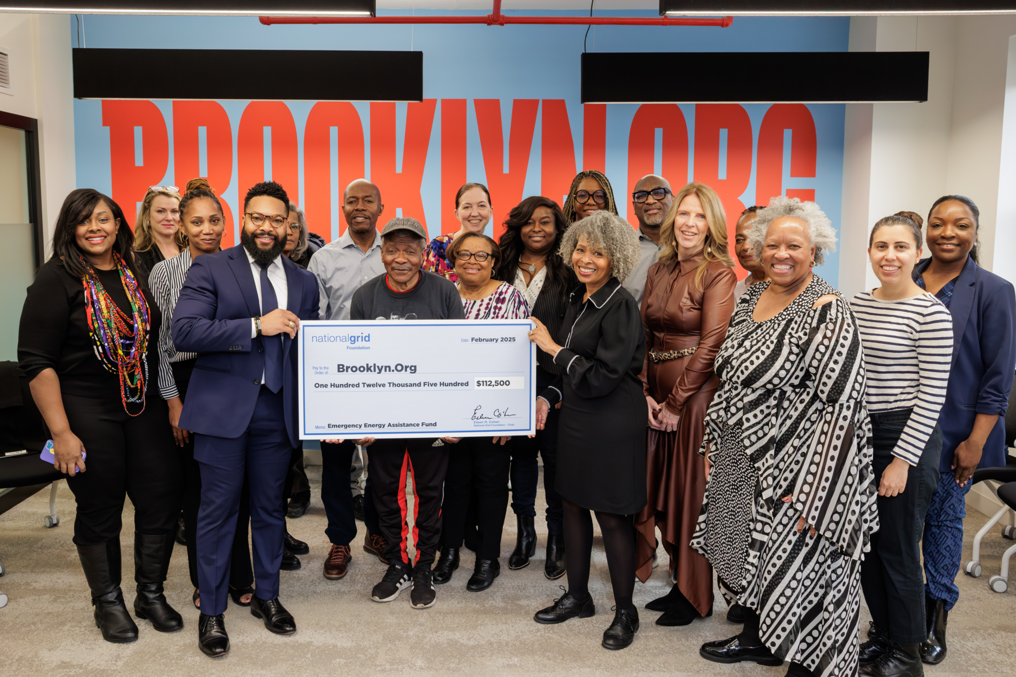 A group of people stand together in an office, holding a large check made out to Brooklyn.Org for $102,000.