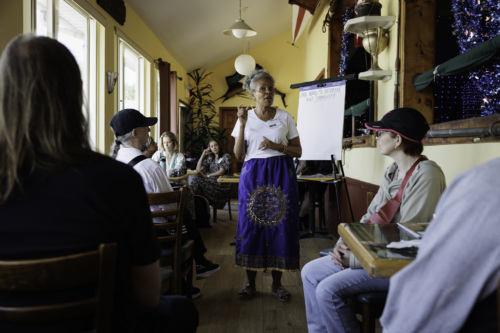 A woman stands speaking to a seated group in a small room with a whiteboard. She wears a white shirt and a blue skirt.