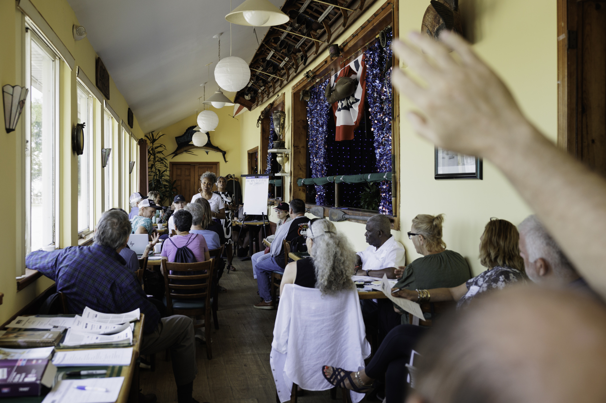 A group of people attending a meeting in a long room with a presenter at the front. One person has their hand raised.