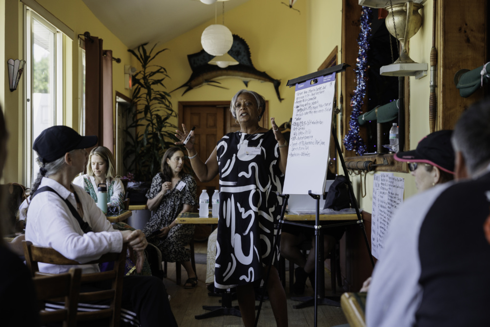 A woman in a patterned dress speaks to a group of people seated around tables in a room, using a flip chart for notes.